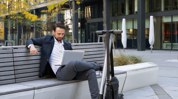 Mann sitzt entspannt auf einer Bank, schaut auf seinen Laptop und hat einen E-Scooter vor sich.