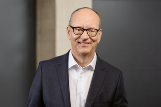 Portrait de Roger Baillod, en costume noir et chemise blanche, devant un mur noir et gris.