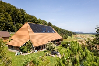 Das Bild zeigt einen grossen Bauernhof mit einer PV-Anlage auf dem Dach. Der Bauernhof befindet sich in einer ländlichen Umgebung im Grünen. Links des Hauses beginnt der Wald.