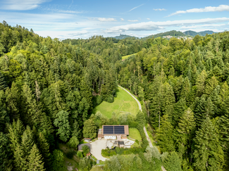 Ein Bauernhaus mit einer PV-Anlage auf dem Dach. Rundherum ist Wald. Ein Waldweg führt zum Haus.