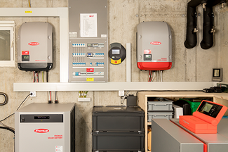 Local technique abritant plusieurs équipements fixés au mur en béton, dont deux onduleurs Fronius, un coffret de protection et de distribution, un système de stockage par batterie, ainsi que des câbles, des conduites et des tuyauteries apparents ; l'installation est claire et réalisée de manière professionnelle.