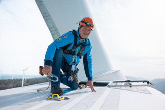 Mann mit hellblauer Jacke und orangefarbenem Sicherheitshelm kniet auf weisser Windturbine und sichert sich mit einem Karabinerhaken