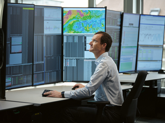 A man sits at a desk and looks at several large screens with figures and price curves.