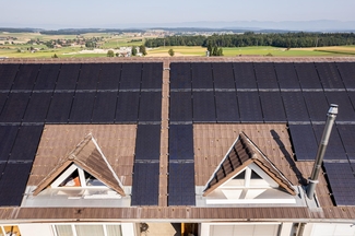 L'image montre le toit à pignon d'un immeuble d'habitation vu de côté. Autour des deux lucarnes, le toit est recouvert d'installations photovoltaïques.