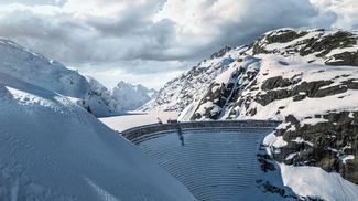 Das Bild zeigt die Grimsel-Staumauer im Winter. Alles ist schneebedeckt und die Sonne scheint zwischen den Wolken durch.