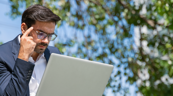 Mann sitzt draussen unter einem Baum und arbeitet konzentriert am Laptop.